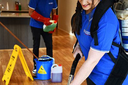 A cleaner selecting the right cleaning product for floor mopping, while the other takes care of vacuuming the floors